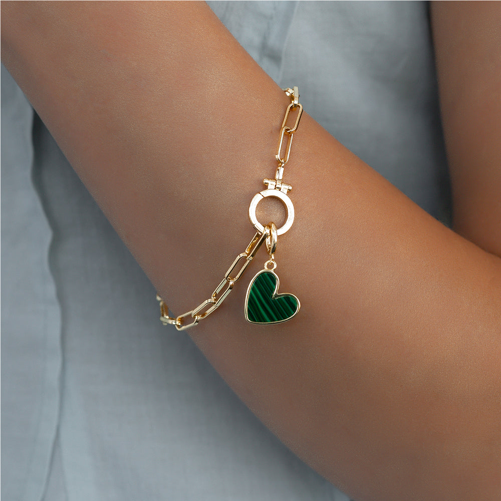 Gold bracelet with a green heart charm on a wrist against a light gray background