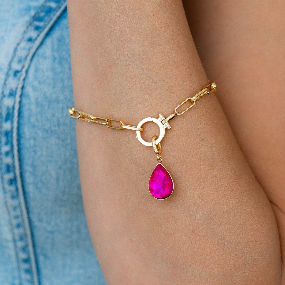 Gold bracelet with a pink teardrop charm on a wrist against a blue denim background