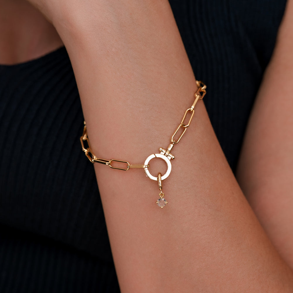 Gold chain bracelet with a small round pendant on a wrist against a dark background