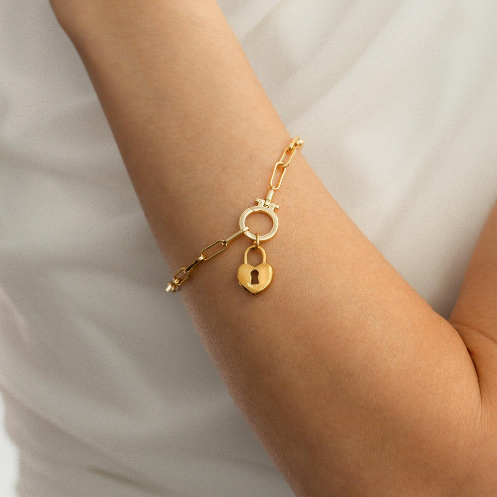 Gold bracelet with a heart-shaped lock charm on a wrist against a neutral background