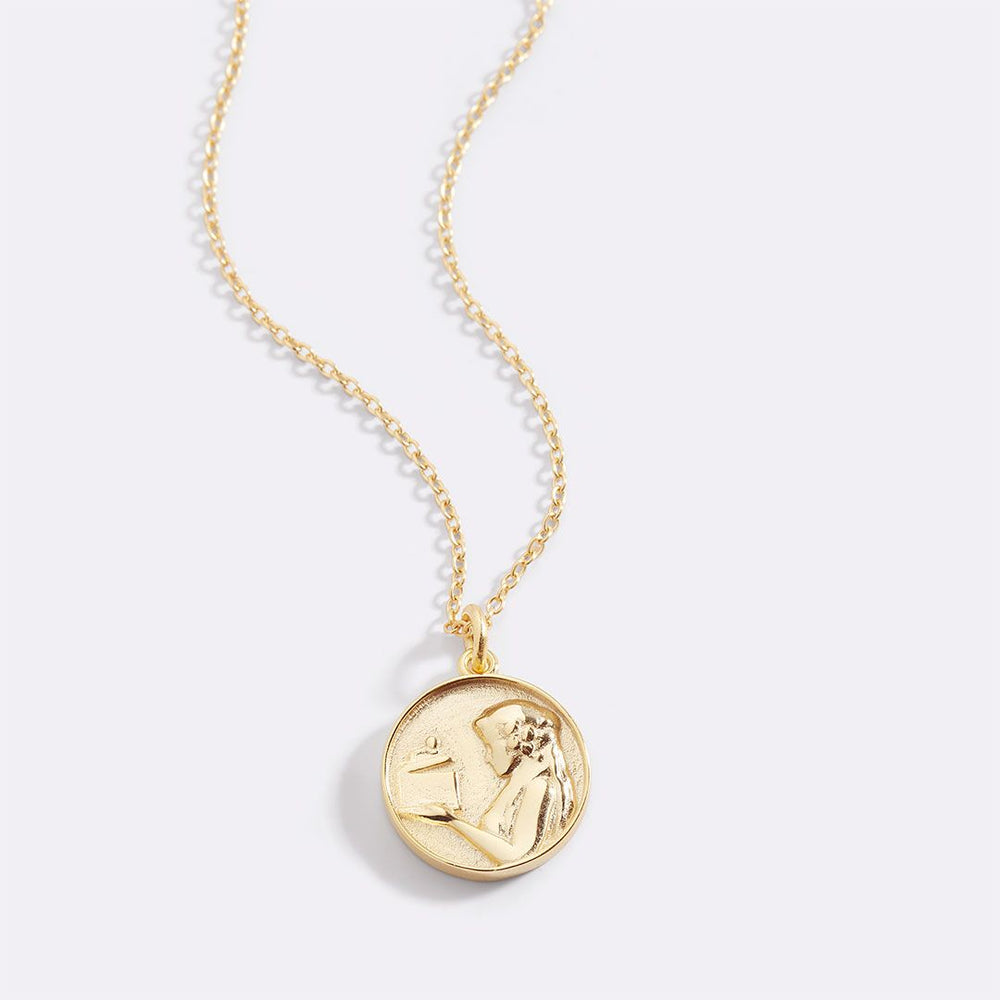 A gold-colored Greek goddess pendant necklace with a detailed circular pendant featuring a face and intricate designs, suspended from a matching gold chain.