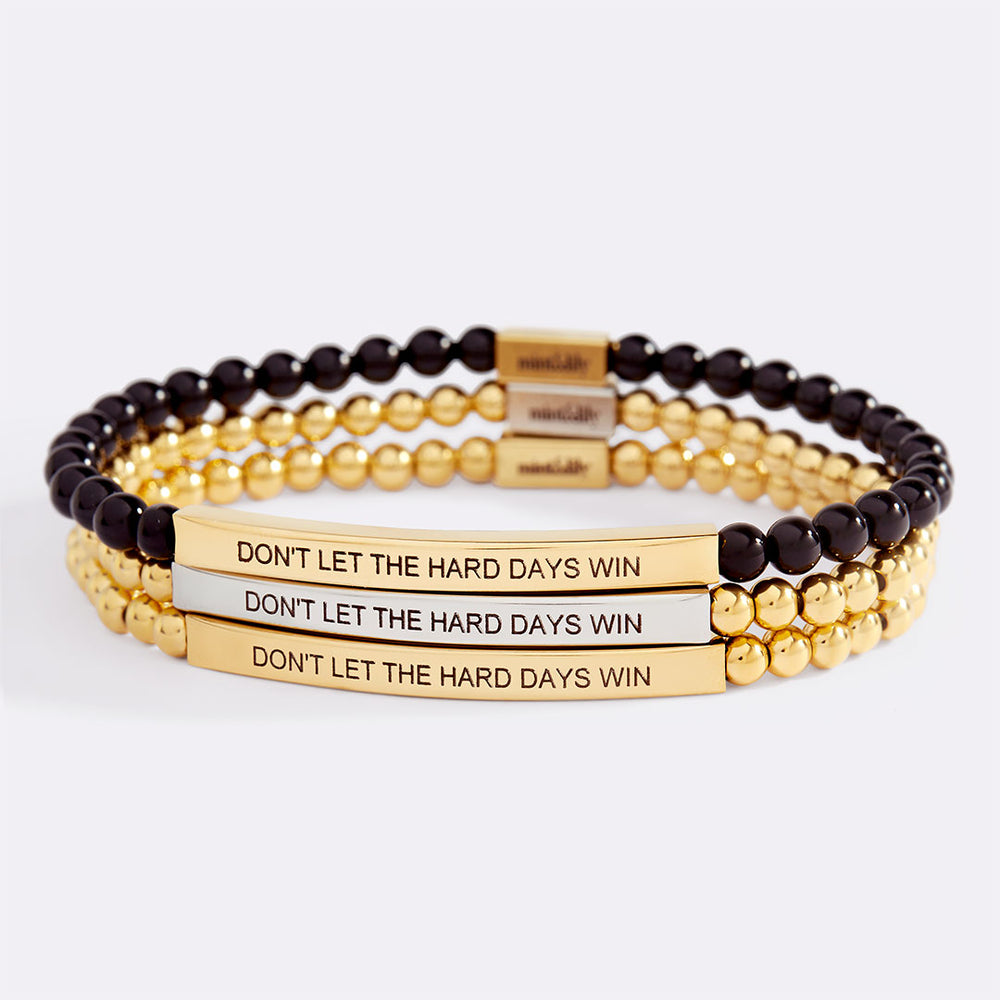 Stack of gold and black beaded bracelets with motivational message on a light gray background
