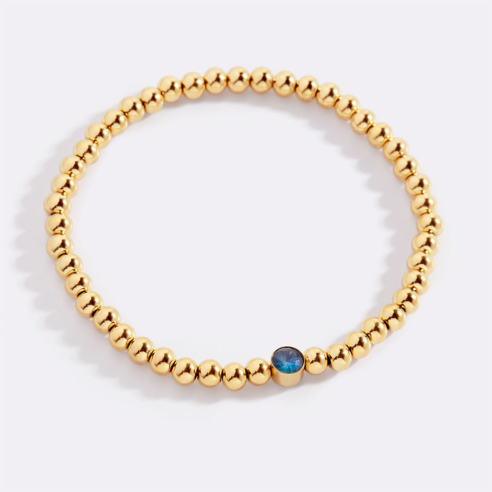 Gold beaded bracelet with a blue stone on a white background