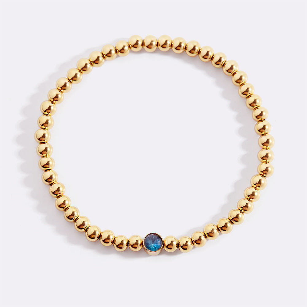 Gold beaded bracelet with a blue stone on a white background