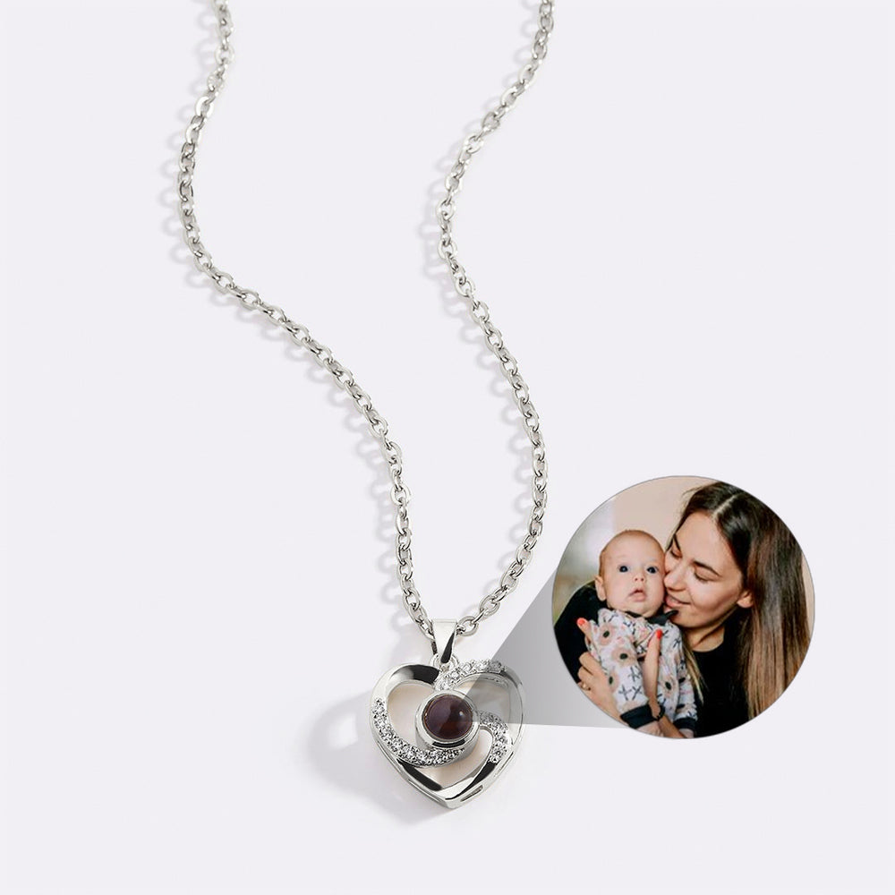 A gold-tone heart-shaped necklace with a pave setting featuring a photo projection stone at the center, displayed alongside an inset image of a woman holding a baby.