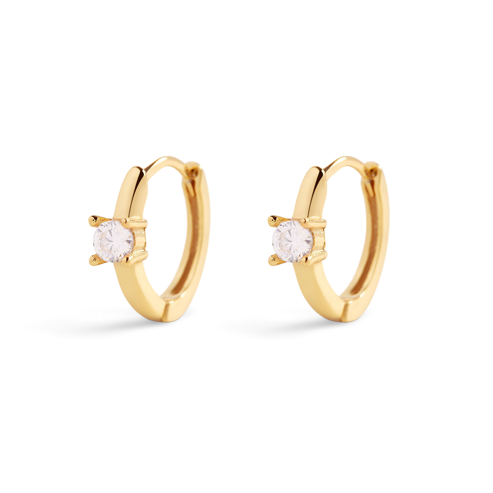 A pair of gold huggie hoop earrings with a single cubic zirconia stone in a 4-prong setting, displayed against a black background.