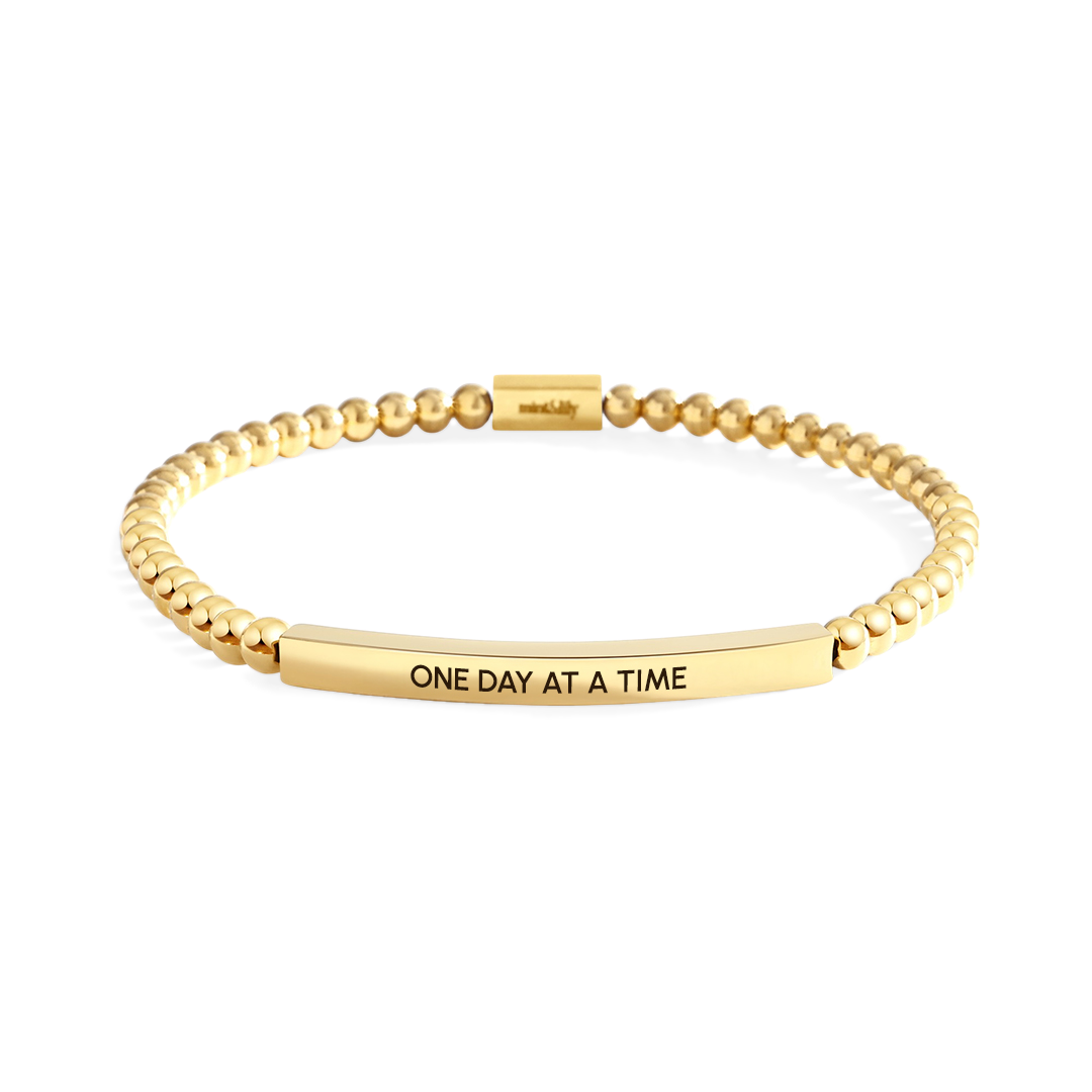 Gold bracelet with 'ONE DAY AT A TIME' engraving on a light gray background