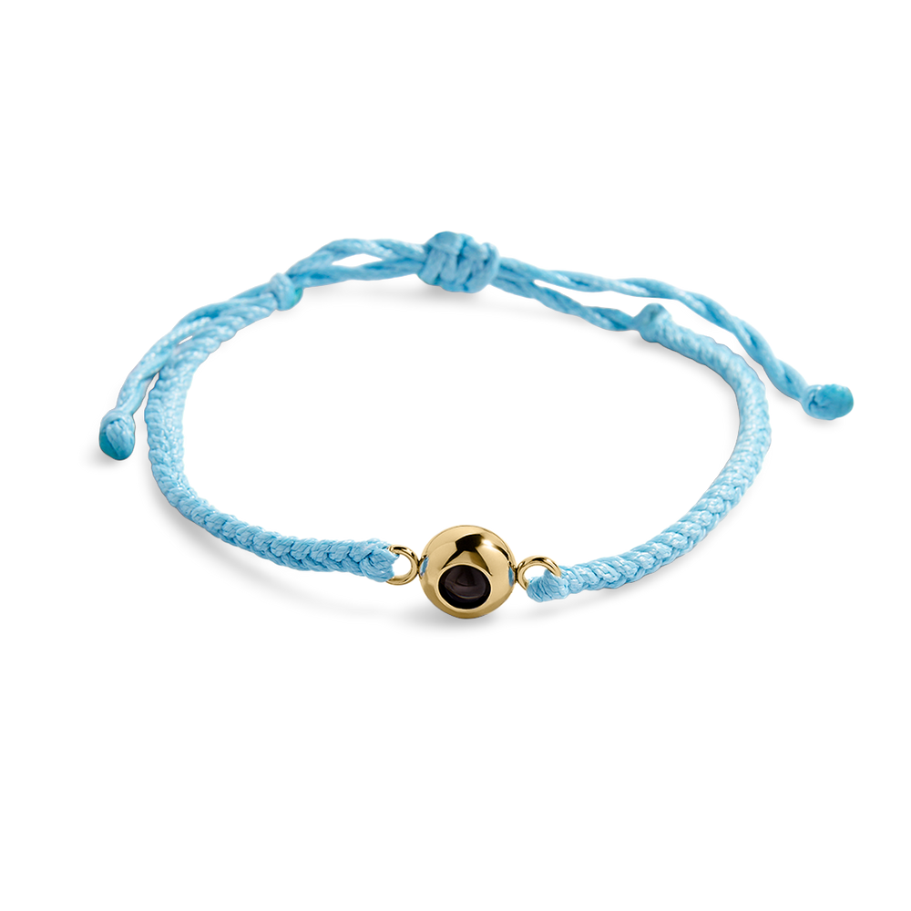 Adjustable blue braided nylon bracelet with a gold-colored circular photo charm in the center, shown with a reflective background.