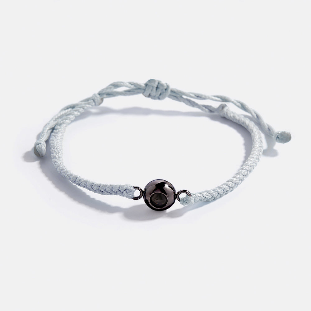 Adjustable blue braided nylon bracelet with a gold-colored circular photo charm in the center, shown with a reflective background.