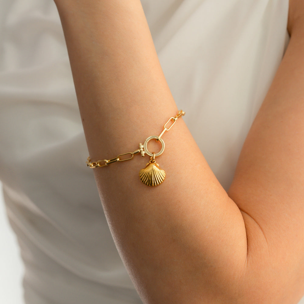 Gold bracelet with a shell charm on a person's wrist against a neutral background