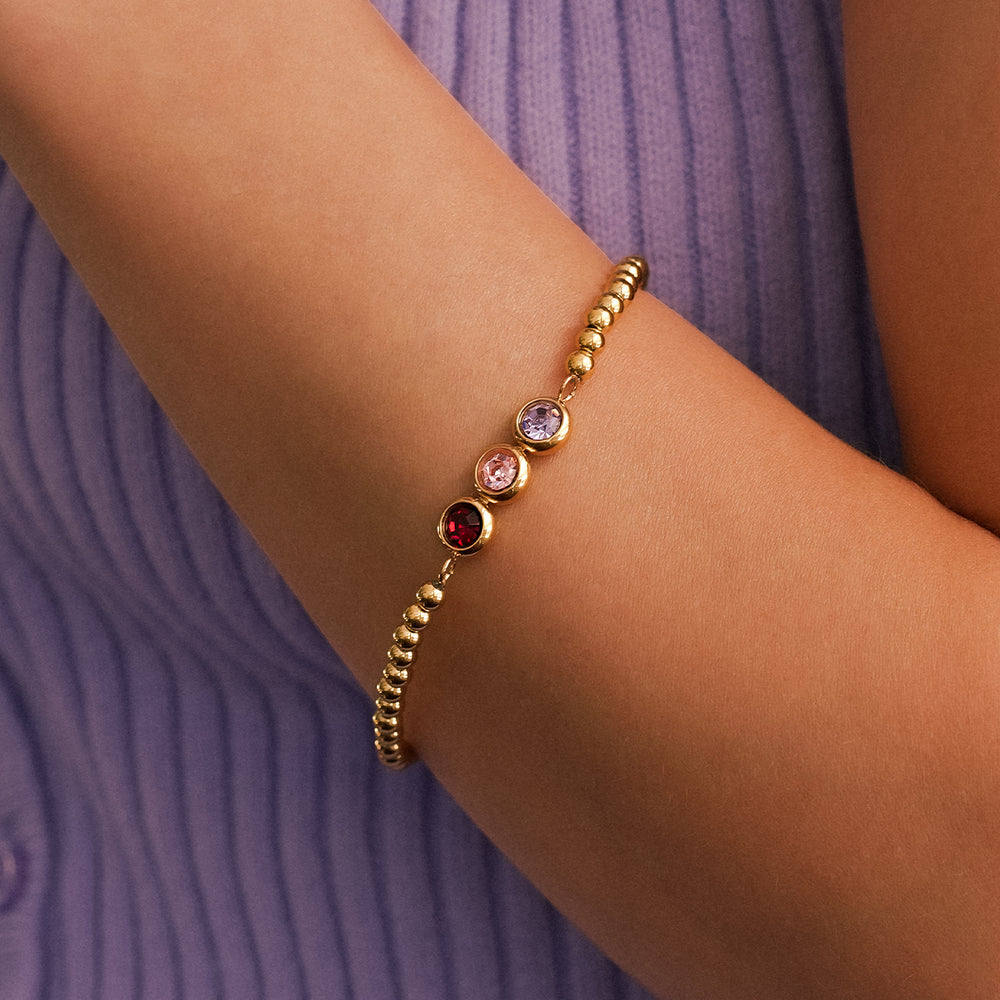 Gold bracelet with colored beads on a wrist against a purple background