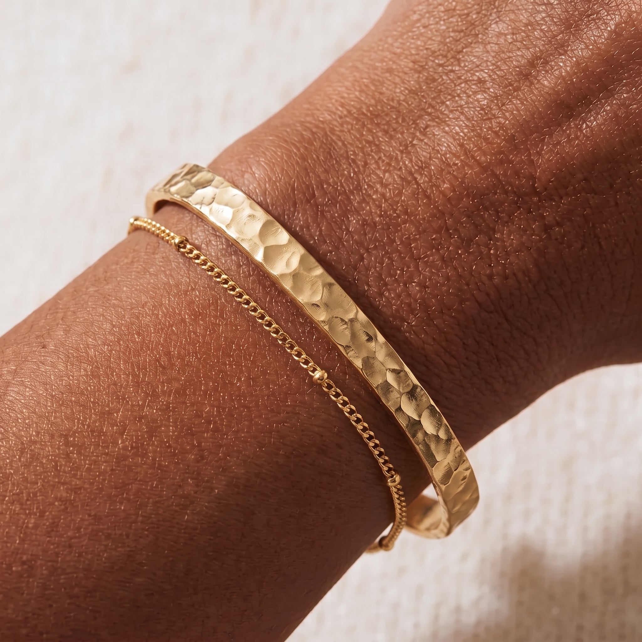 Hammered gold cuff bracelet stacked with a delicate gold satellite chain bracelet on a wrist against a soft cream linen background.