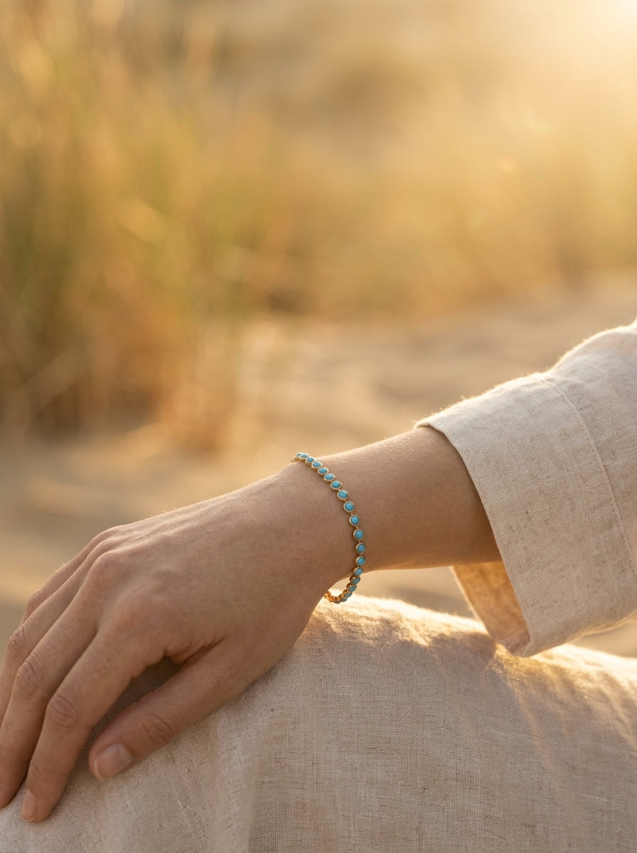 A woman's wrist wearing the gold Turquoise Bezel-Set Tennis Bracelet, resting on her linen skirt in low golden-hour sunlight beside sunlit dune grass.