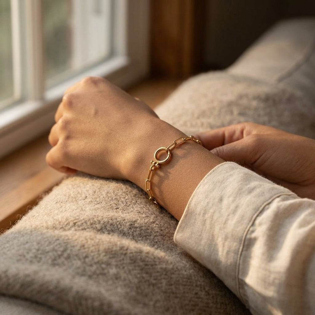 A wrist wearing the gold Mint Paperclip Bracelet resting on a soft oatmeal wool throw in warm low light from a nearby window.