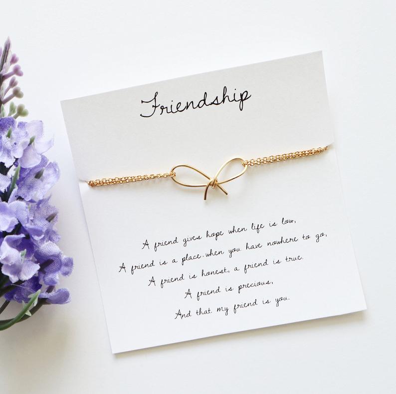 A gold-colored friendship bracelet with a bow detail, presented on a card with the word 'Friendship' written on it, accompanied by a quote about friendship. The bracelet is displayed next to purple flowers.